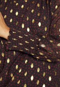 Tessuto nero con motivi di foglie bordeaux e oro; presenta polsini arricciati con texture per un aspetto aderente; elegante e leggero.