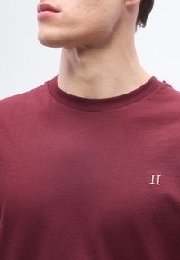 Νέος άντρας που φοράει μπλουζάκι μπόρντο με στρογγυλή λαιμόκοψη και λευκή κεντητή επιγραφή "II" στο στήθος, με το κεφάλι γυρισμένο στο πλάι.