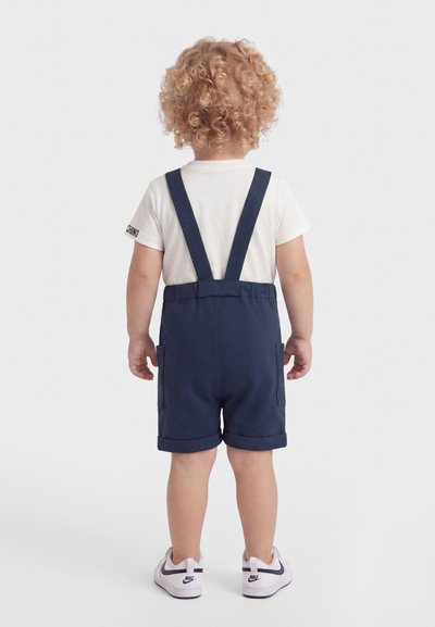 Toddler with curly blonde hair wearing a white t-shirt, navy blue shorts with suspenders, and white Nike sneakers, standing facing away.