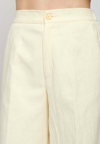 Pantaloni crema a vita alta con chiusura a bottone singolo in vita, indossati da una persona non visibile.