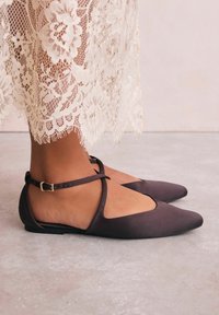 Ballerines en cuir marron avec un bout pointu, présentant un design découpé, une bride de cheville avec boucle, et une texture lisse. Ourlet de robe en dentelle visible.