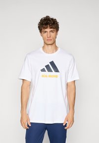 Hvid bomulds t-shirt med et sort geometrisk logo og "REAL MADRID" i gul skrift, korte ærmer og rund halsudskæring.