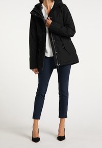 Parka noire à capuche avec fermeture éclair, poches latérales et cordons ajustables. Portée sur un haut blanc et associée à un pantalon bleu marine.