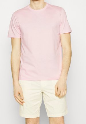 Mężczyzna w gładkim jasnoróżowym T-shircie z krótkim rękawem i kremowych szortach stoi na białym tle.