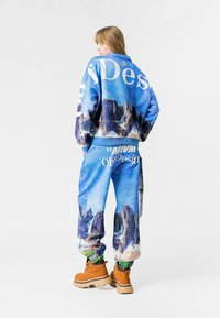 Chándal gráfico azul con imágenes de paisajes y texto grande. Se complementa con botas color camel. El material parece texturizado con puños elásticos.