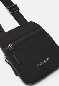 Sacoche noire en bandoulière avec un matériau texturé, poche zippée et logo. La sangle réglable et les accents lisses améliorent la fonctionnalité.