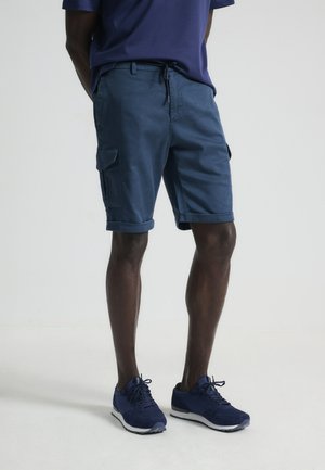 Pantalones cortos de carga en color marino con dobladillos enrollados, fabricados en una mezcla de algodón. Presentan bolsillos laterales y una cintura con cordón, combinados con zapatillas marinas.