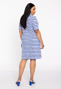 Gestreiftes Kleid in Weiß und Blau, kurze Ärmel, knielang, aus einem strukturierten Stoff. Getragen mit blauen Blockabsatzsandalen.