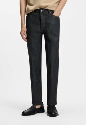 Mann trägt dunkelblaue Jeans mit geradem Bein, beigen Oberteil und schwarze Lederloafer, steht mit einer Hand in der Hosentasche.