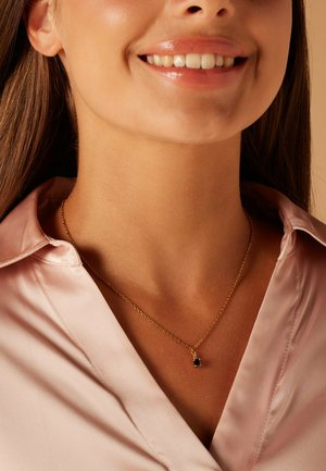 Collier en or avec une chaîne fine et un petit pendentif en pierre précieuse verte, porté sur un chemisier en satin rose clair. Gros plan sur le décolleté.