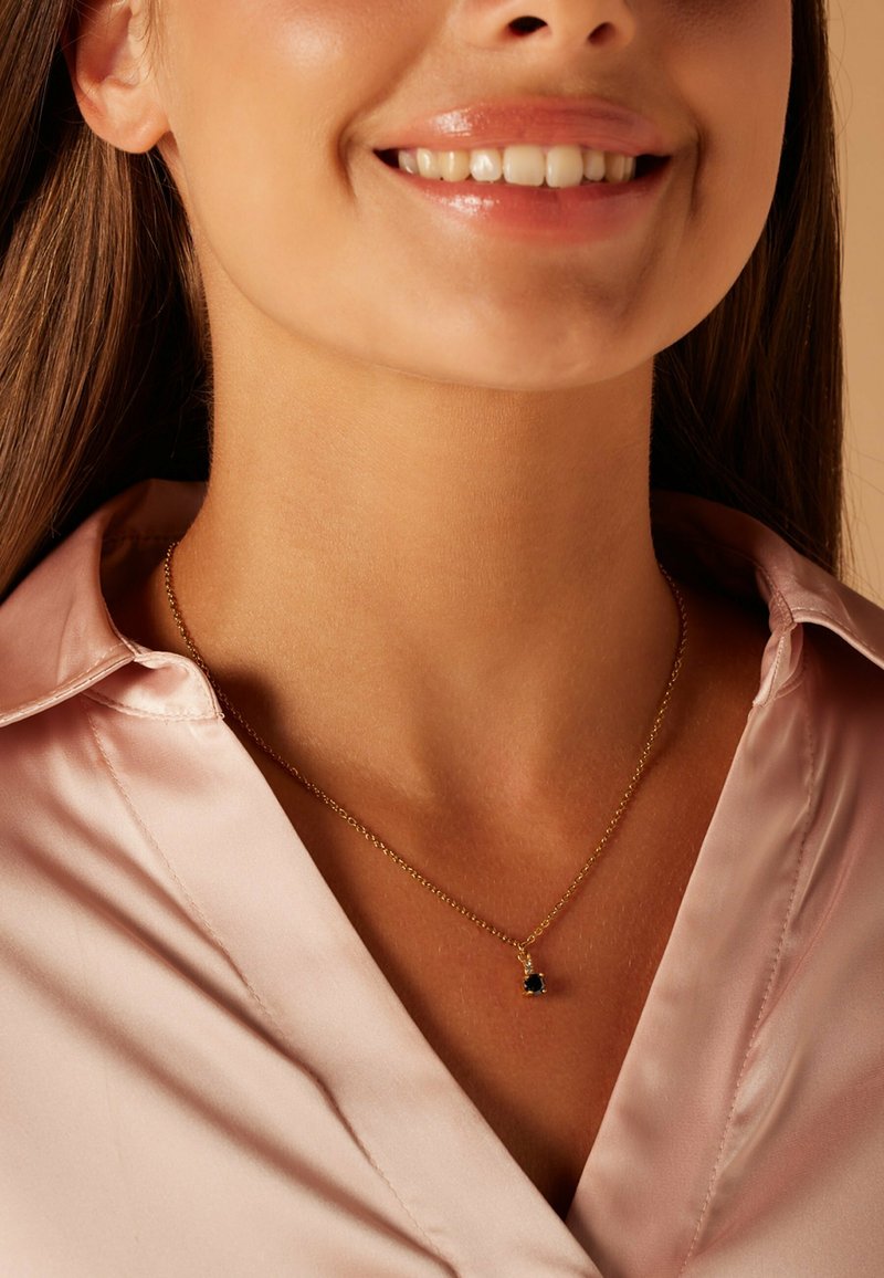 Collier en or avec une chaîne fine et un petit pendentif en pierre précieuse verte, porté sur un chemisier en satin rose clair. Gros plan sur le décolleté.