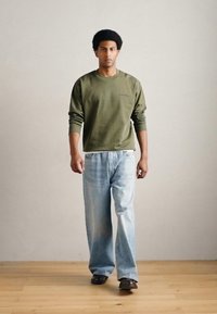 Camisa de manga larga verde oliva, vaqueros holgados azul claro y zapatos oscuros. Modelo con cabello corto y rizado de pie en una habitación de color claro.