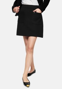 Black textured skirt with a fitted design, featuring two front pockets and a wide waistband. Paired with black flats and a white top.