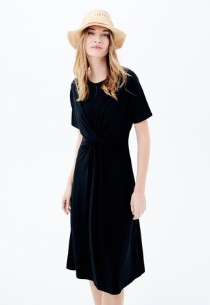 Robe noire jusqu'aux genoux avec un détail de taille torsadé, manches courtes, fabriquée dans un tissu doux et lisse. Complétée par un chapeau en paille.