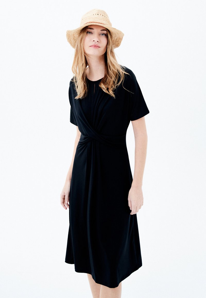 Robe noire jusqu'aux genoux avec un détail de taille torsadé, manches courtes, fabriquée dans un tissu doux et lisse. Complétée par un chapeau en paille.