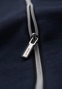 Gros plan d'une fermeture éclair métallique argentée sur un tissu foncé avec un tiret rectangulaire gravé "ombre" en noir et blanc.