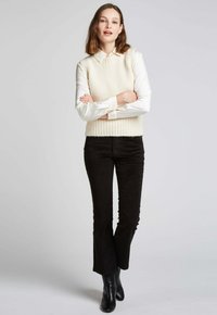 Gilet en tricot crème superposé sur une chemise col blanc, associé à un pantalon en velours côtelé noir évasé et des bottes noires jusqu'aux genoux.