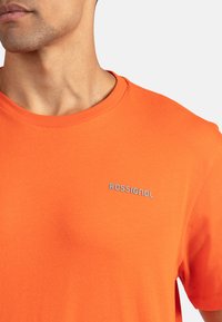 Maglietta arancione a maniche corte con scollatura rotonda, caratterizzata da un logo grigio "ROSSIGNOL" sulla parte sinistra del petto. Materiale in cotone, texture liscia.