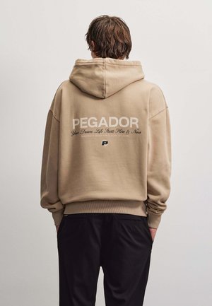 Personne aux cheveux bruns porte un sweat à capuche beige avec "PEGADOR" et un texte au dos, associé à un pantalon noir, sur un fond clair uni.