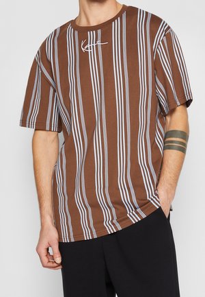 Chemise marron à manches courtes avec des rayures blanches verticales et un logo blanc signature sur la poitrine, portée par une personne avec un pantalon noir et un tatouage sur le bras.