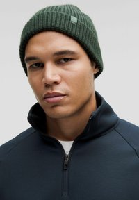 Gorro de punto acanalado verde combinado con una chaqueta de cremallera azul marino, que presenta un alto cuello y un sutil detalle de cremallera. Forma ajustada, sin patrones.