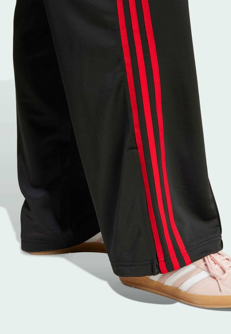 Pantalones de chándal negros con rayas rojas a los lados, con un corte relajado. Combinados con zapatillas rosa claro con detalles en blanco y suela beige.