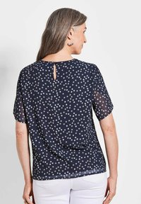 Navyblauwe doorschijnende blouse met witte stippen, korte mouwen, sleutelhalsdetail aan de achterkant en een losse pasvorm. Structuurstof met een lichte uitstraling.