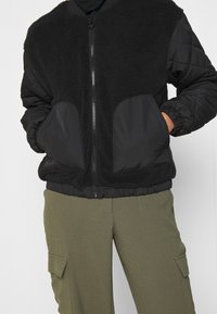 Veste bomber noire avec un haut en polaire et des manches matelassées noires. Dotée d'une fermeture éclair et d'une ceinture élastique. Associée à un pantalon cargo olive.