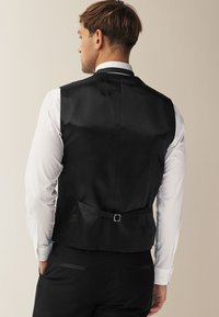 Chaleco de satén negro con un corte a medida, que presenta un detalle de hebilla en la parte posterior y se lleva sobre una camisa blanca de manga larga.