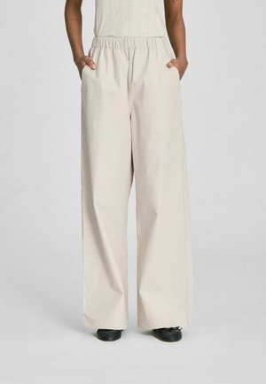 Osoba nosiaca béžové nohavice s vysokým pásom a širokými nohavicami, ruky v kapsách, čierne balerínky na nohách, na jednoduchom pozadí.