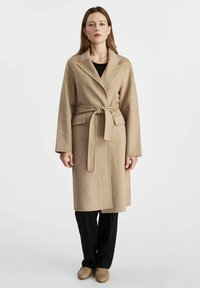 Beżowy wełniany płaszcz z paskiem, z wyłogami, kieszeniami bocznymi i długimi rękawami, noszony na czarnych spodniach i butach wsuwanych. Gładkie tło.