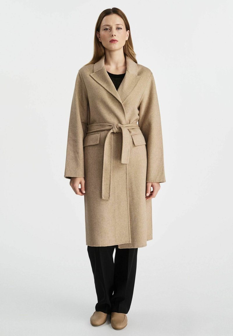 Beżowy wełniany płaszcz z paskiem, z wyłogami, kieszeniami bocznymi i długimi rękawami, noszony na czarnych spodniach i butach wsuwanych. Gładkie tło.