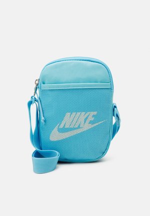 Malá modrá taška Nike přes rameno s nastavitelným popruhem a bílým logem na přední síťované kapse na jednobarevném pozadí.