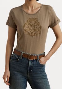 Beige bomulls t-shirt med korta uppvikta ärmar och en strukturerad guldig broderad design på framsidan, ihop med blå denimjeans och ett brunt bälte.