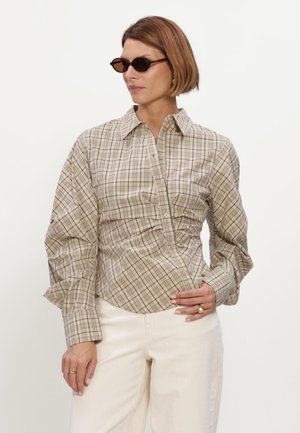 Vrouw met kort bruin haar, die schildpadkleurige zonnebril draagt, een beige geruit overhemd met lange mouwen en off-white broek, staand met één hand op de heup.