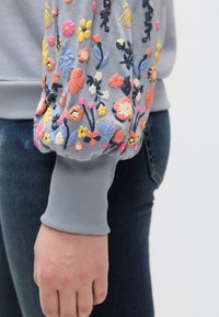 Ανοιχτό γκρι φούτερ με πολύχρωμες κεντητές ανθοδέσμες στα φουσκωτά μανίκια; ριμπ μανσέτες; εφαρμοστή σιλουέτα συνδυασμένη με σκοτεινό τζιν.