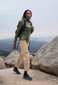 Grønn zip-up hettegenser, beige cargo-bukser og grå turstøvler. Scenen har steinete terreng og en fjellbakgrunn.