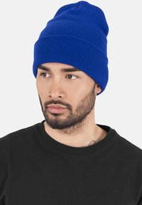 Căciulă tricotată albastră cu o textură ribbată și o margine rimbată. Pur tată cu un tricou negru cu guler rotund, având mâneci scurte și un finisaj neted.