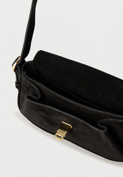 Μαύρη σουέντ τσάντα ώμου με χρυσό κούμπωμα, ανοιχτό πάνω μέρος και ρυθμιζόμενο λουράκι, εμφανίζεται μπροστά σε απλό ανοιχτόχρωμο φόντο.