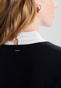 Mörkblå tröja med vit krage. Har en knappstängning bak och strukturerad stickad tyg. Röd örhängeaccent synlig.