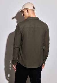 Olivově zelená košile s dlouhým rukávem a knoflíkovými manžetami, texturovaná tkanina a zakřivený lem. Nosit s lehkým béžovým kloboukem. Model otočen zády.