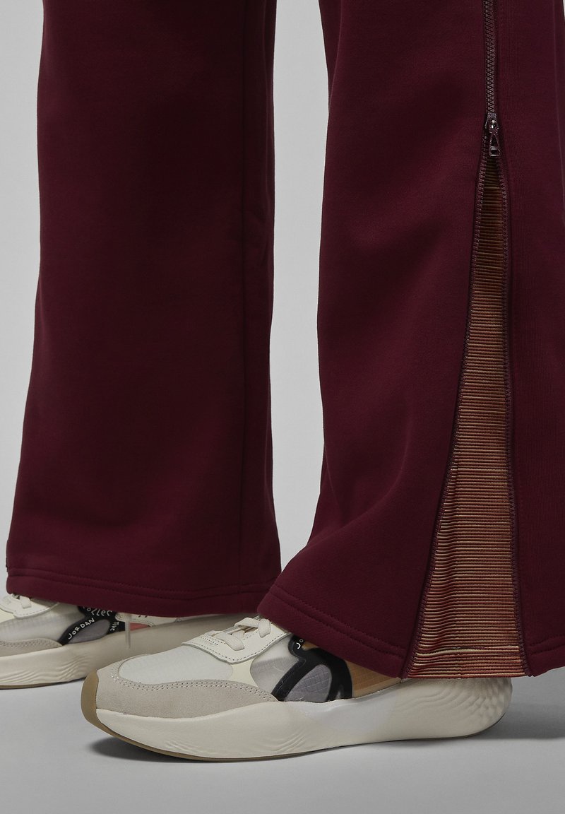 Sneaker mit beigen, weißen und schwarzen Details, getragen zu burgunderfarbenen Hosen mit seitlichen Reißverschlüssen, die teilweise geöffnet sind und ribbeligen Stoff zeigen.