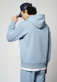 Sudadera azul claro hecha de una tela suave, con capucha y puños acanalados. El modelo lleva una gorra azul marino y blanca, vista desde detrás.