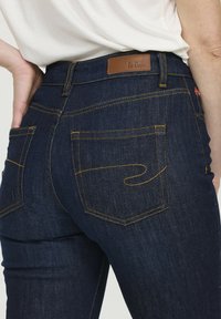 Jean en denim bleu marine avec une coupe ajustée, présentant des coutures jaunes, deux poches arrière et une étiquette en cuir marron à la taille.