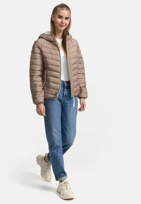 Beige gewatteerde jas met capuchon, heeft een gewatteerd ontwerp en een zilveren rits; gedragen met blauwe jeans en lichtgekleurde sneakers.