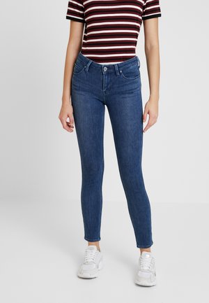 Femme portant un jean skinny bleu foncé, des baskets blanches et un t-shirt à manches courtes à rayures horizontales bordeaux, blanches et noires, debout.