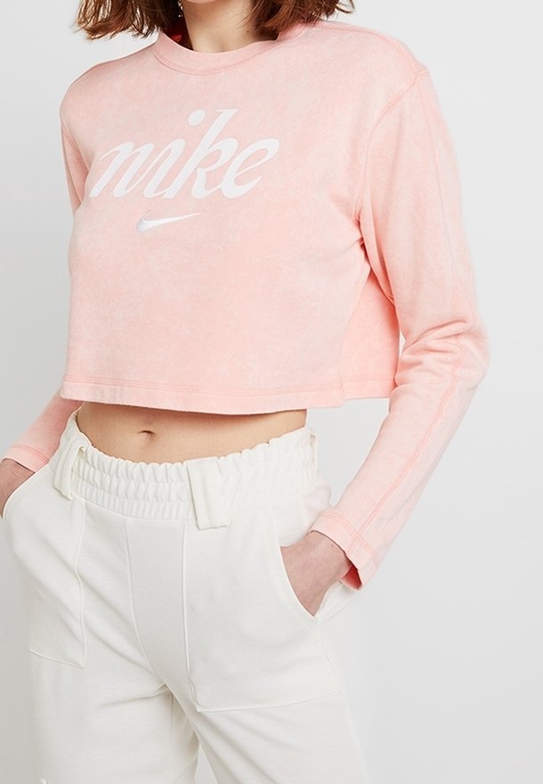 Avskuren rosa sweatshirt med vit "nike"-logotyp, tillverkad av mjukt tyg. Har långa ärmar och en avslappnad passform, kombinerad med vita byxor.