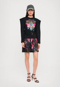 Camisa negra de manga larga con bordado floral colorido, combinada con una falda plisada negra y floral. Accesorios incluyen una gorra gris y tacones negros.