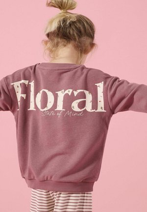 Barn med lyst hår i en knold bærer en mauve sweatshirts med teksten "Floral State of Mind" på ryggen imod en pink baggrund.