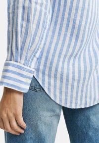 Camicia di lino a righe blu e bianche con polsino abbottonato, orlo curvo e maniche arrotolate, indossata con jeans di denim blu chiaro.